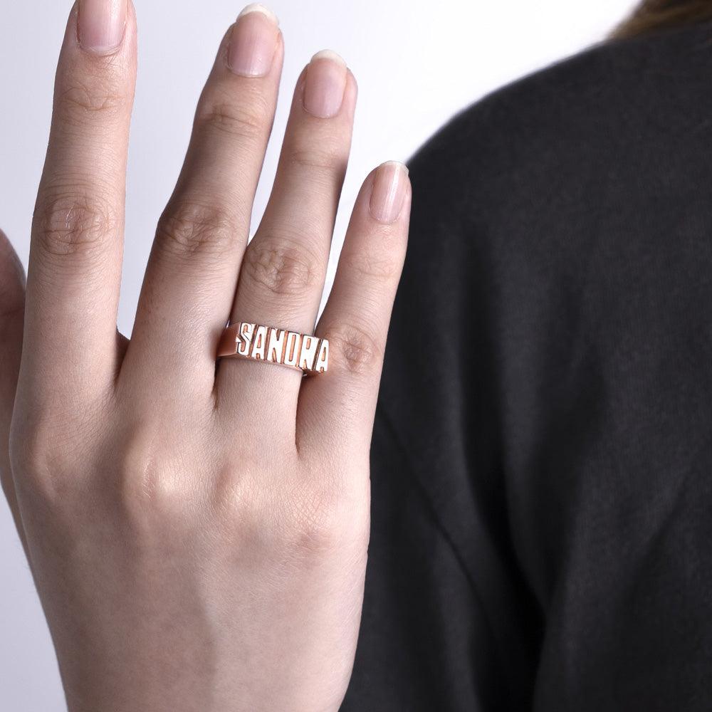 A person wearing a rose gold custom name ring with the name 'Sandra,' showcasing the ring on their finger against a dark shirt.