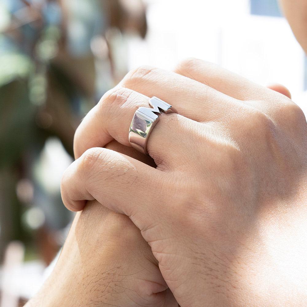 A person wearing a silver custom name ring with the initial 'M,' showcasing the ring on their finger with hands clasped together in a natural light setting.