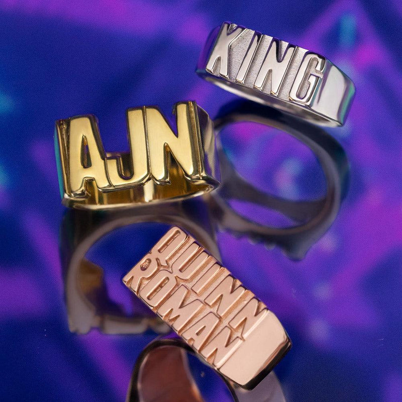 Three custom name rings in gold, silver, and rose gold, featuring the names 'AJN,' 'KING,' and 'QUINN ROMAN' displayed against a vibrant purple background.