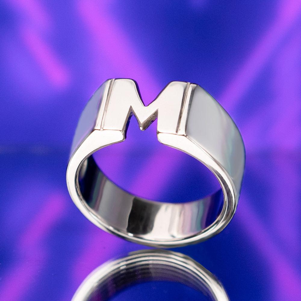 A silver custom name ring featuring the initial 'M,' displayed against a vibrant purple background with reflective surface.