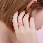 A close-up of a person wearing a rose gold ring with the name "Amelia" written in cursive, featuring a red gemstone set at the end of one of its bands, on their finger near their ear.