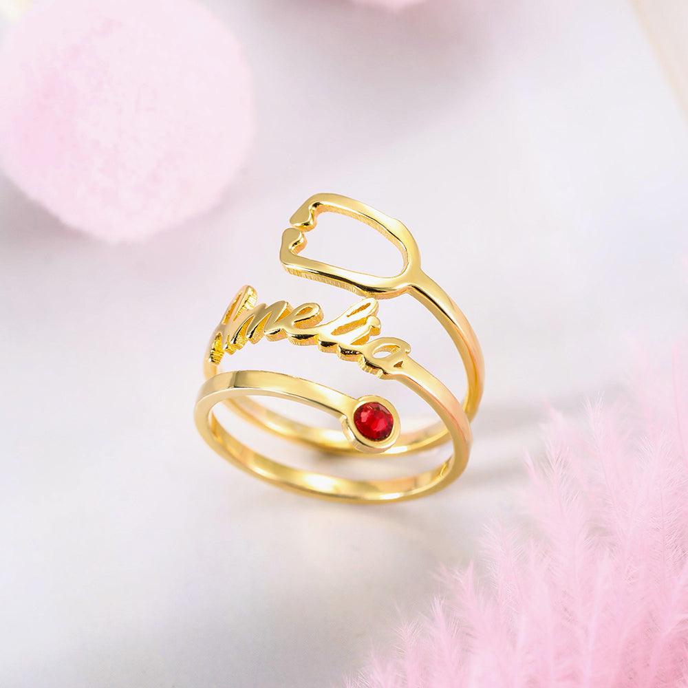 A gold ring with the name "Amelia" written in cursive, featuring a red gemstone set in the middle of one of its bands, surrounded by pink decorations.