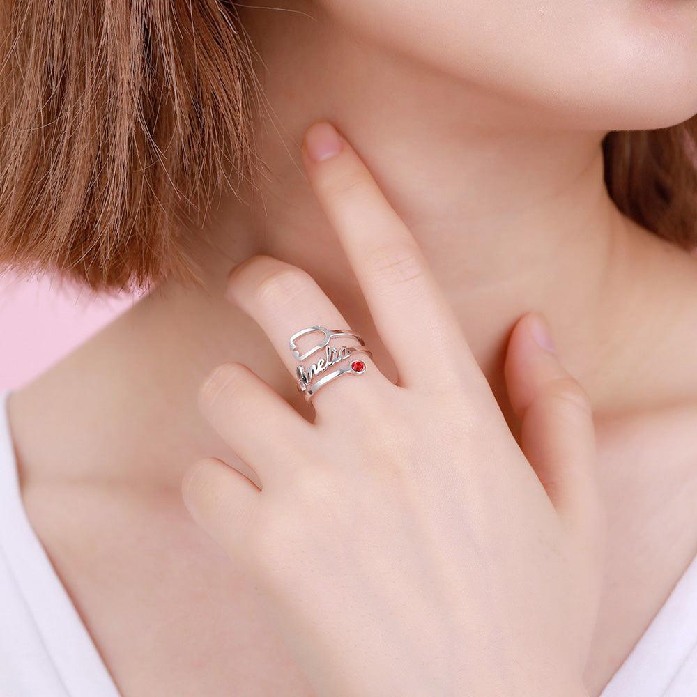 A close-up of a person wearing a silver ring with the name "Amelia" written in cursive, featuring a red gemstone, on their finger, with their hand near their neck.