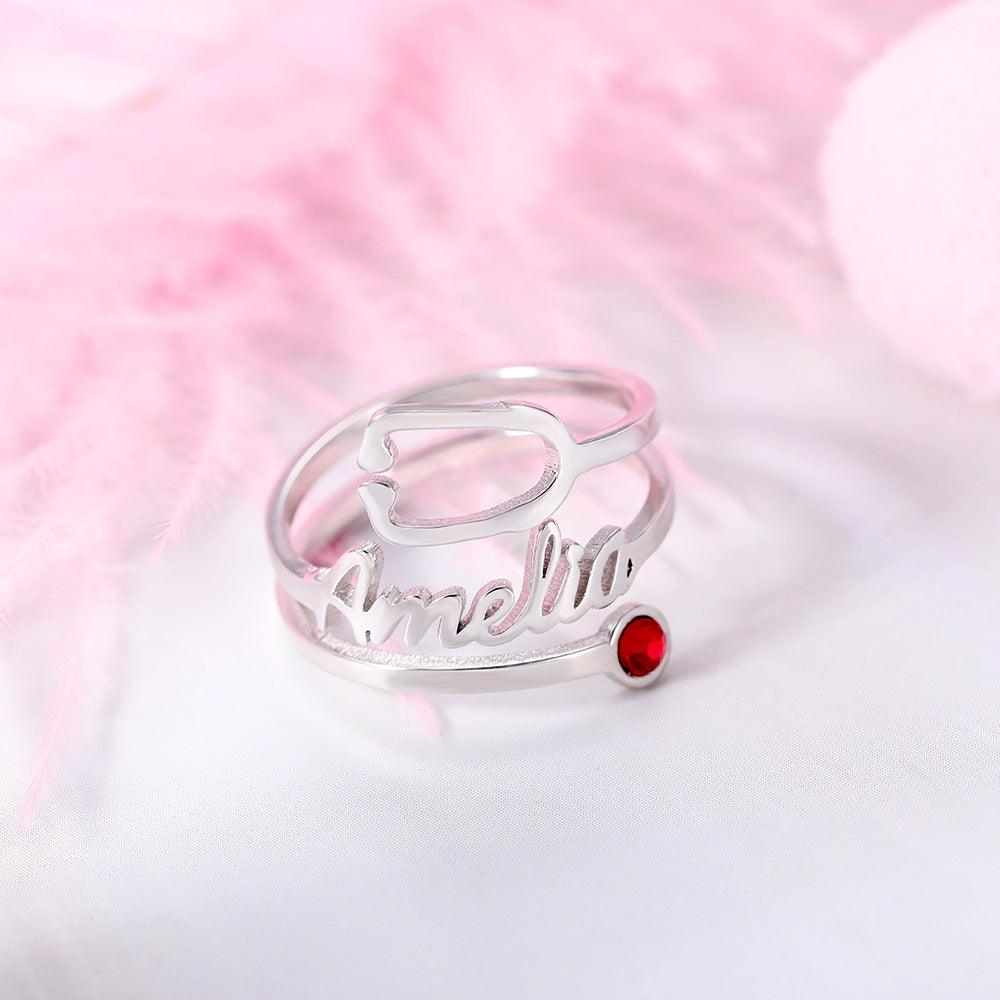 A silver ring with the name "Amelia" written in cursive, featuring a red gemstone, placed on a white surface with pink feather decorations in the background.