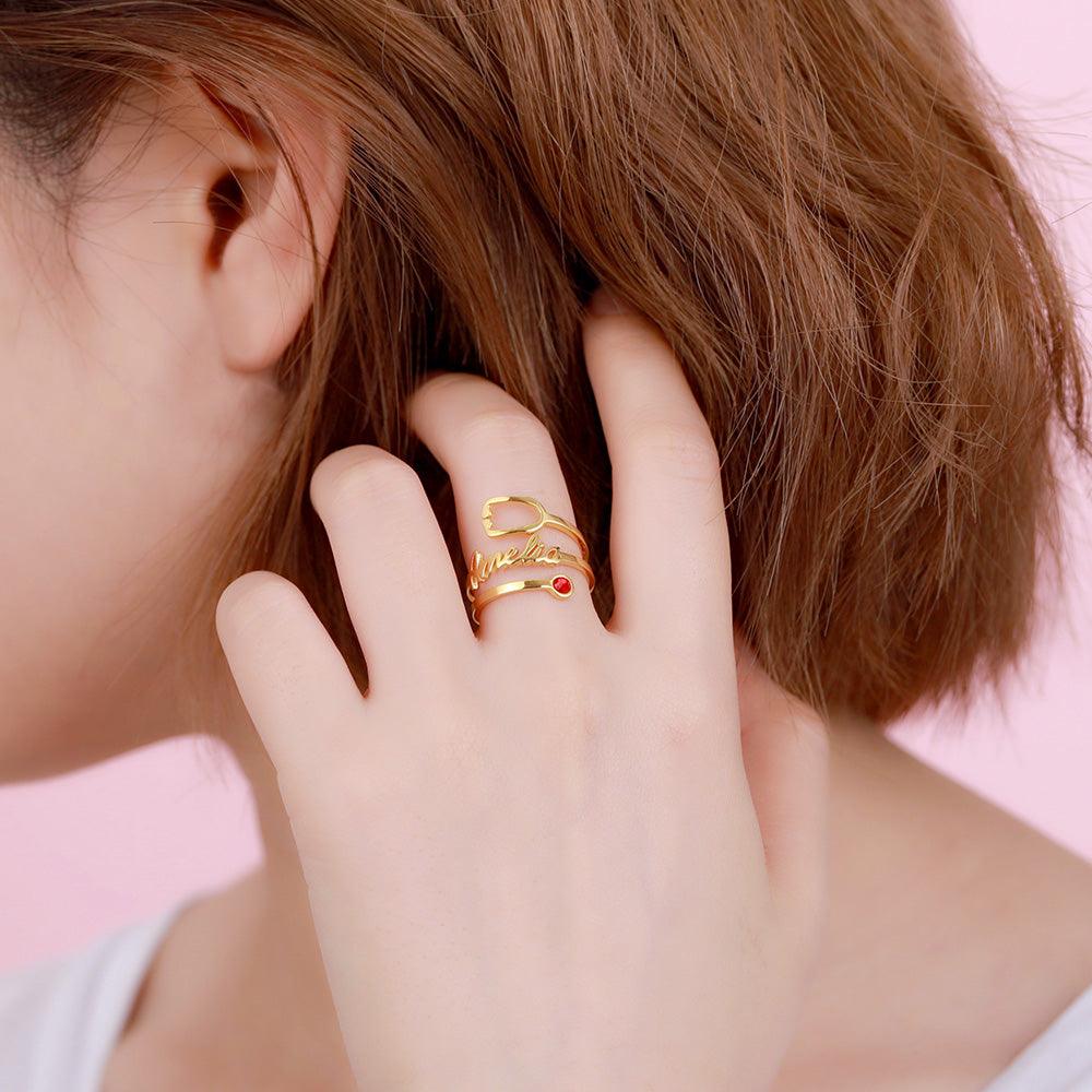 A close-up of a person wearing a gold ring with the name "Amelia" written in cursive, featuring a red gemstone set at the end of one of its bands, on their finger near their hair.