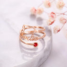 A rose gold ring with the name "Amelia" written in cursive, featuring a red gemstone set at the end of one of its bands, placed on a marble surface with dried flowers in the background.