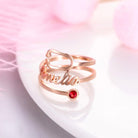 A rose gold ring with the name "Amelia" written in cursive, featuring a red gemstone set at the end of one of its bands, placed on a white surface with pink decorations.