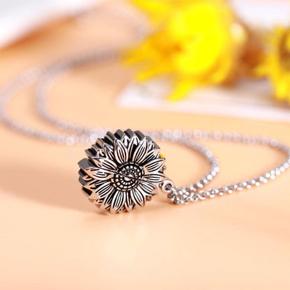 A silver sunflower-shaped locket necklace closed, with intricate petal detailing, displayed against a soft peach background with a blurred yellow flower in the back.