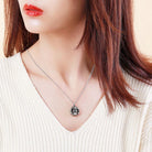 A woman wearing a silver sunflower-shaped locket necklace against a white V-neck sweater, with her red hair framing her face and a neutral background.