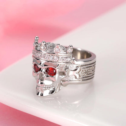 "Silver skull ring with crown, red eye gems, and intricate engravings on a pink backdrop.