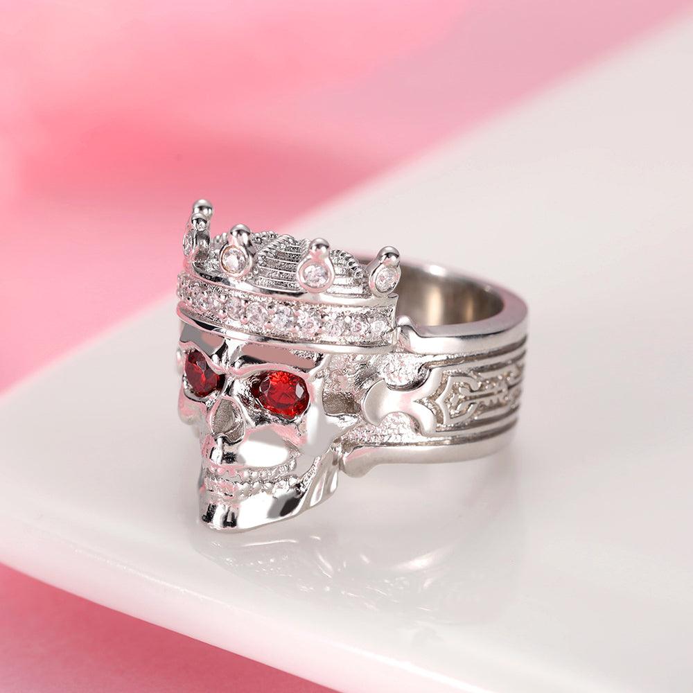 "Silver skull ring with crown, red eye gems, and intricate engravings on a pink backdrop.