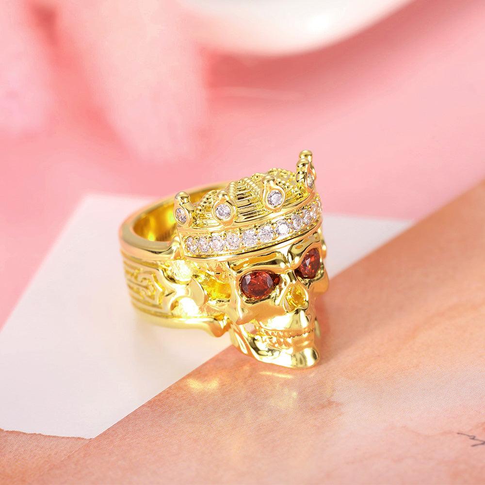 Golden skull ring with sparkling crown and red eyes on a soft pink background.