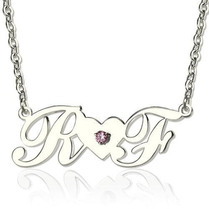 A silver necklace with cursive initials "R" and "F" connected by a heart-shaped bridge with a pink gemstone in the center.