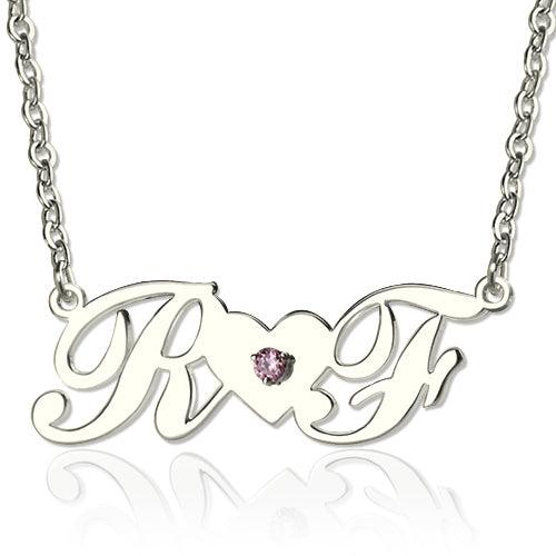 A silver necklace with cursive initials "R" and "F" connected by a heart-shaped bridge with a pink gemstone in the center.