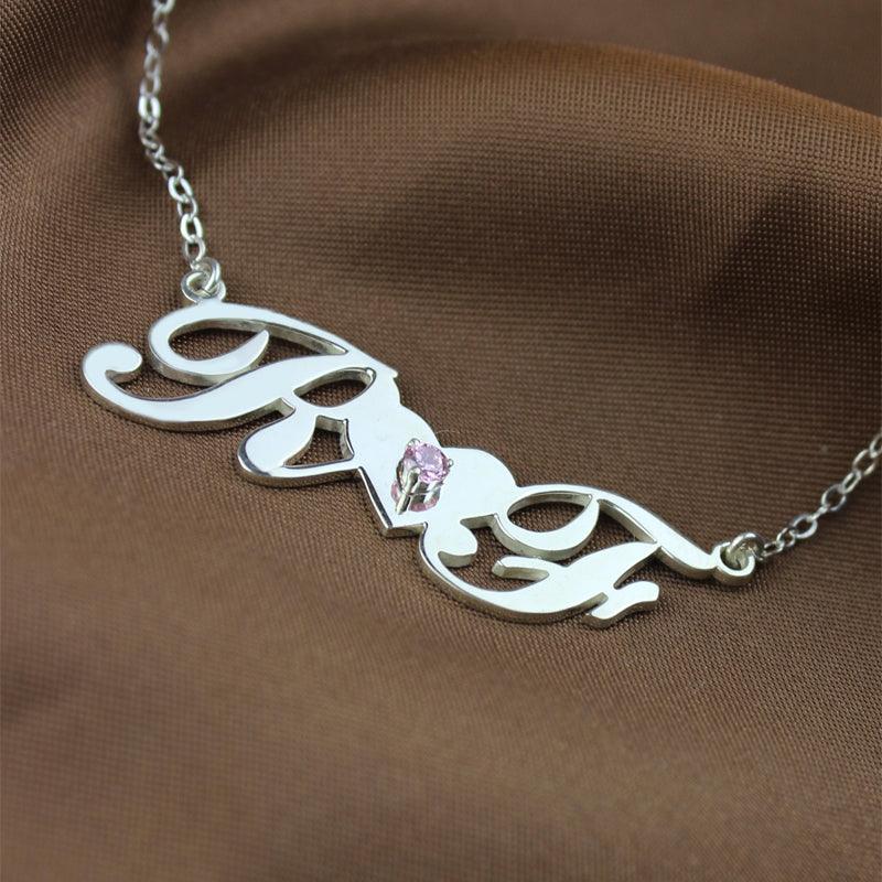 A silver necklace with cursive initials "R" and "F" connected by a heart-shaped bridge with a pink gemstone in the center, displayed on a brown fabric background.