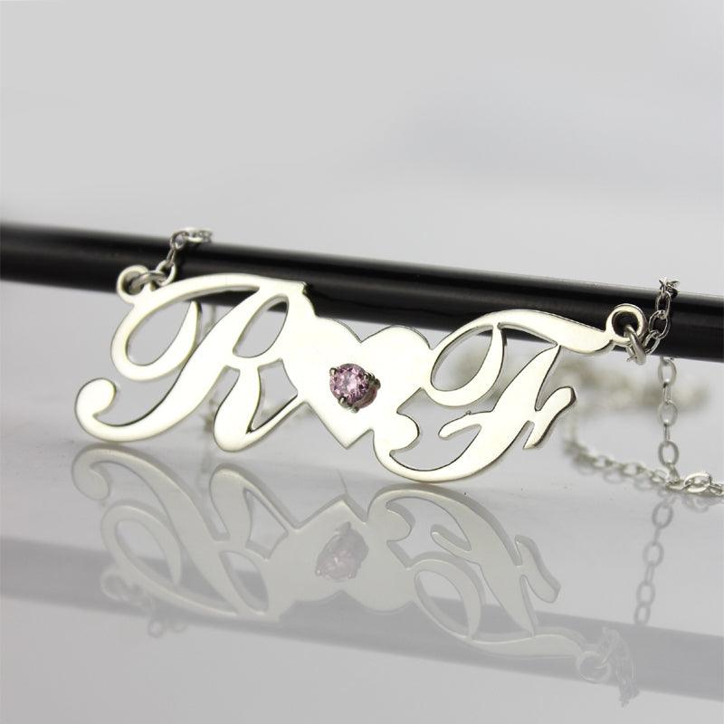 A silver necklace with cursive initials "R" and "F" connected by a heart-shaped bridge with a pink gemstone in the center, displayed on a reflective surface.