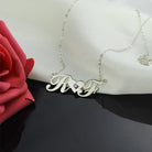 A silver necklace with cursive initials "R" and "F" connected by a heart-shaped bridge with a pink gemstone in the center, displayed on a white fabric with a red rose nearby.