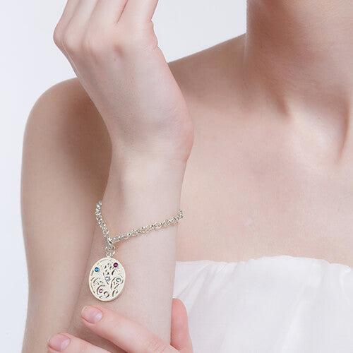 A woman in a white strapless top wearing a silver charm bracelet with a tree pendant featuring colored gemstones, touching her arm lightly with her fingers.