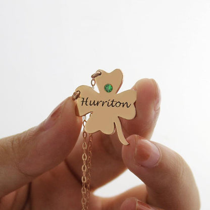 A hand holding a rose gold necklace with a four-leaf clover pendant inscribed with "Huririton" and a green gemstone embedded in one of the leaves.