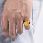 Close-up of a man's hand wearing a golden signet ring with a family crest, against a denim background.