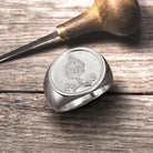 Silver signet ring with embossed family crest on wooden surface, next to an engraving tool.