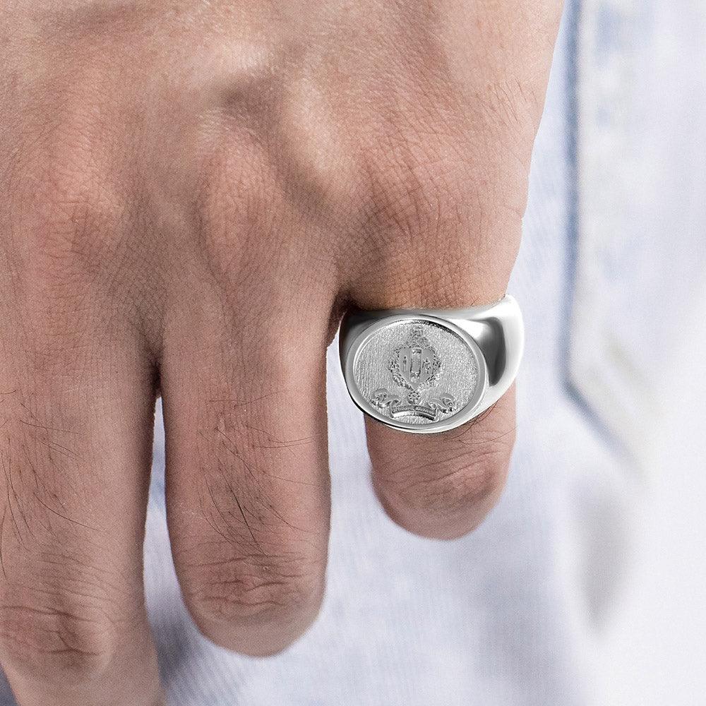 Close-up of a man's hand wearing a silver signet ring with a detailed crest, against a light fabric background.