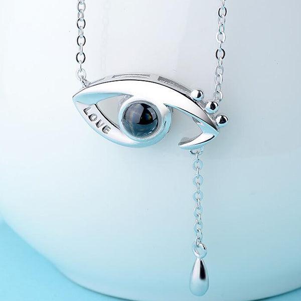 A close-up of a silver eye-shaped pendant necklace with the word "LOVE" engraved, featuring a dangling chain, set against a light background.