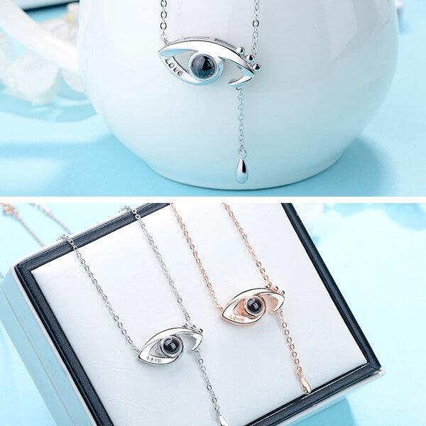 Two images: the top shows a silver eye-shaped pendant necklace with "LOVE" engraved, dangling from a white vase; the bottom shows both silver and rose gold versions displayed in a box.