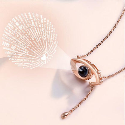 A rose gold eye-shaped pendant necklace projecting a beam of light that forms a heart surrounded by tiny text, showcasing a unique and intricate design.