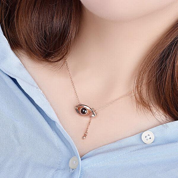 A close-up of a woman wearing a rose gold necklace featuring an eye-shaped pendant with a dangling chain, set against a light blue shirt.